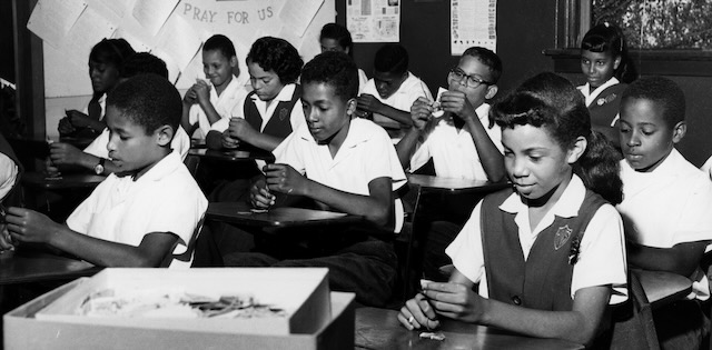 A group of young students in a classroom holding stamps at their desks.