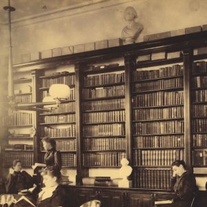 An old sepia toned photo of students studying in a library in front tall bookshelves.