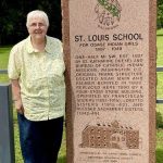 A woman with short white hair and wire glasses is wearing a butter-yellow collared button-up shirt and dark wash jeans. She's smiling for a picture on a sunny day while standing next to a stone plaque that says 'ST. LOUIS SCHOOL' and additional information.