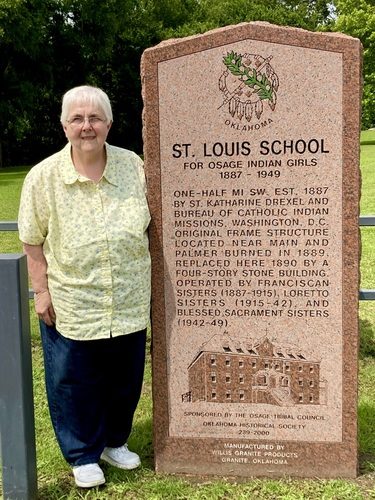 A woman with short white hair and wire glasses is wearing a butter-yellow collared button-up shirt and dark wash jeans. She's smiling for a picture on a sunny day while standing next to a stone plaque that says 'ST. LOUIS SCHOOL' and additional information.