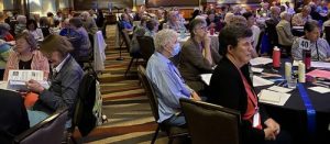 A wide view of a filled event room with people sitting at round tables and looking towards the right. On their tables they have blue ribbon, black table cloths, a number, candles, and assorted papers. Most of the attendees are wearing lanyards and dress-clothes.