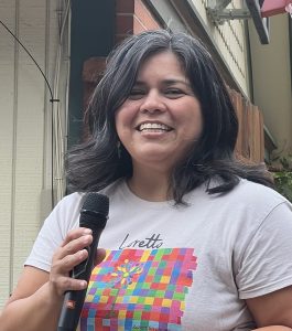 A woman with shoulder length dark brown hair smiling brightly while holding a microphone and wearing a white t-shirt with a colorful checkered graphic on the front.