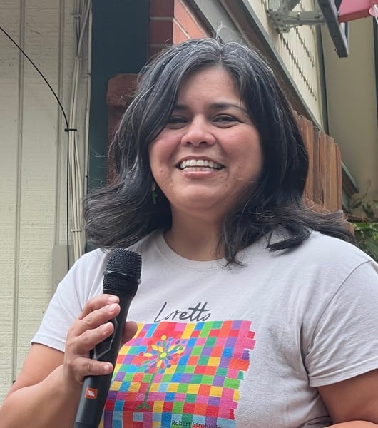 A woman with shoulder length dark brown hair smiling brightly while holding a microphone and wearing a white t-shirt with a colorful checkered graphic on the front.