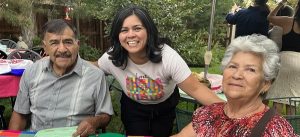 A man on the left, and a woman on the right sitting together at a table, with a woman leaning down in between them to smile for a picture together. They are outdoors in front of greenery and spruce trees with other tables and people behind them.