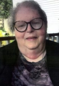 A woman with short, dark grey hair, and big, dark-framed glasses smiling for a headshot picture wearing a black cardigan and a dark grey, patterned blouse.