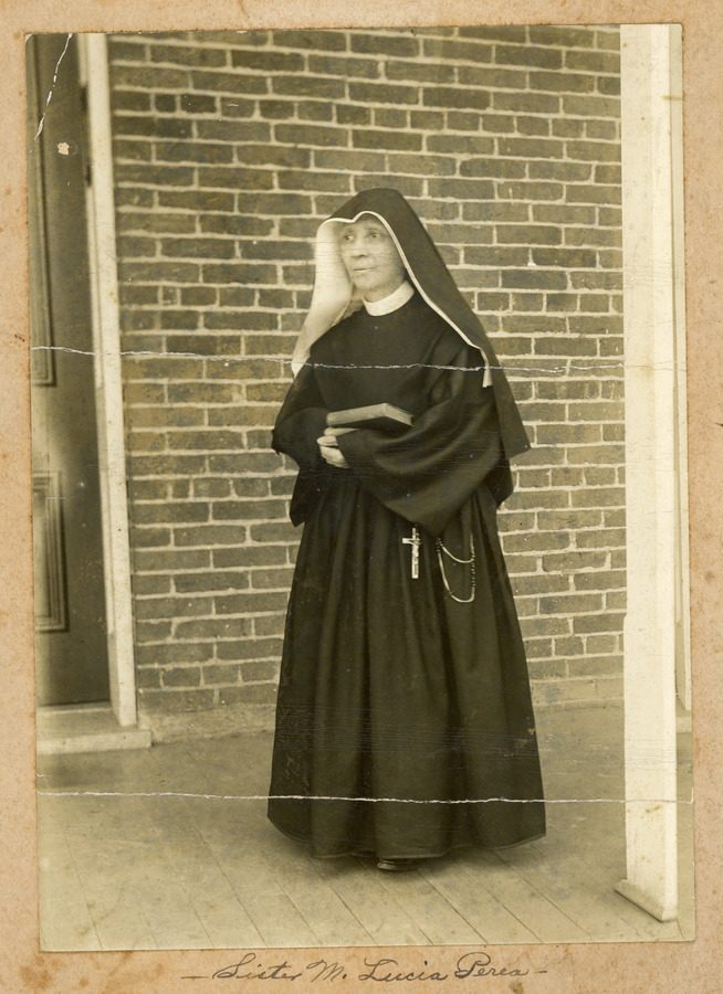 Archival photo of an older Sister Lucia Perea in habit and holding a book standing on the porch of a brick building.