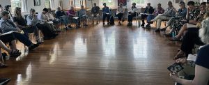 A large room with wooden floors and big bright windows with a large crescent of chairs with people sitting and holding papers in their laps, talking together in a group discussion.