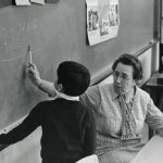 Historical photo of an older teacher, sitting in a chair, working with a young boy to solve the math math problem written on the blackboard behind them.