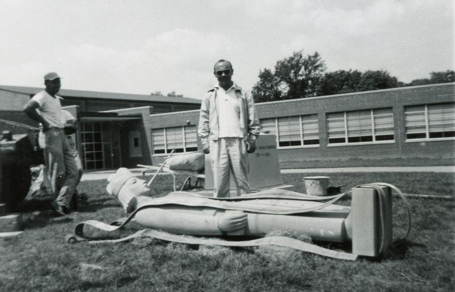 Rudy Torrini stands over a large stone statue laying on the ground. Heavy straps lay about the statue and a workman stands off to one side.