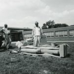 Rudy Torrini stands over a large stone statue laying on the ground. Heavy straps lay about the statue and a workman stands off to one side.
