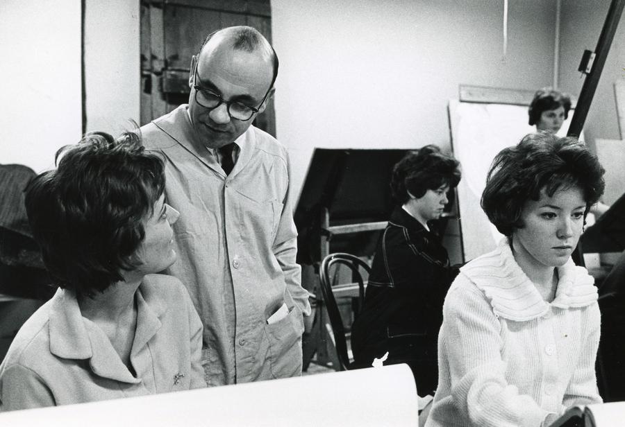A man with glasses and a coat talks with a female student in this archival photo of a drawing class.