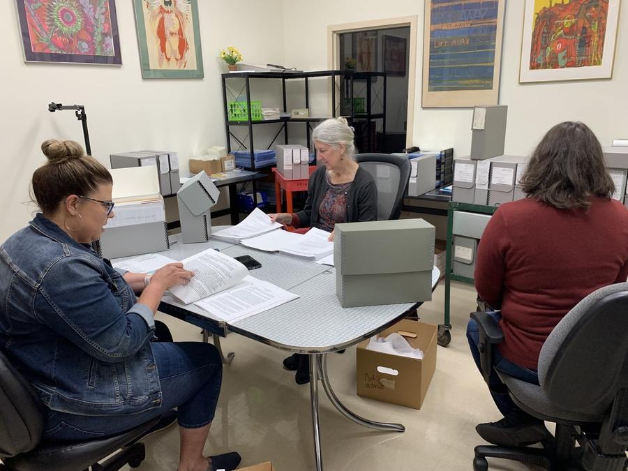 Three women sit in an office meeting room sorting papers.