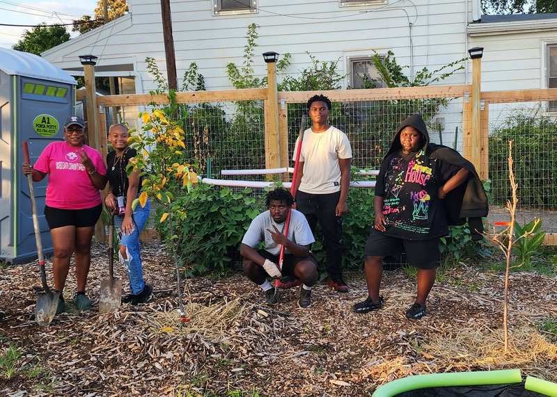 Five people holding garden tools pose for a photo with two newly planted trees in a community garden.