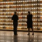Two women admiring a grand wall of back-lit glass jars with assorted dirt and pebbles. It is a memorial site, each jar has the ground material from different lynching sites, putting the sheer volume of the tragedies into perspective.