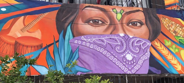 A bright colorful mural of oranges and blues shows a hispanic woman's face partially covered by a purple bandana