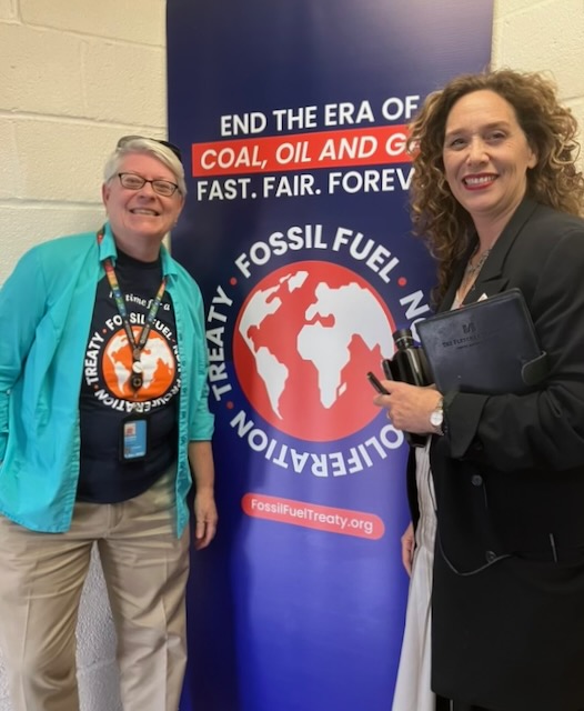 Two women smiling in front of a banner with the world on it that says "End the Era of Coal, Oil and Gas. Fast. Fair. Forever."