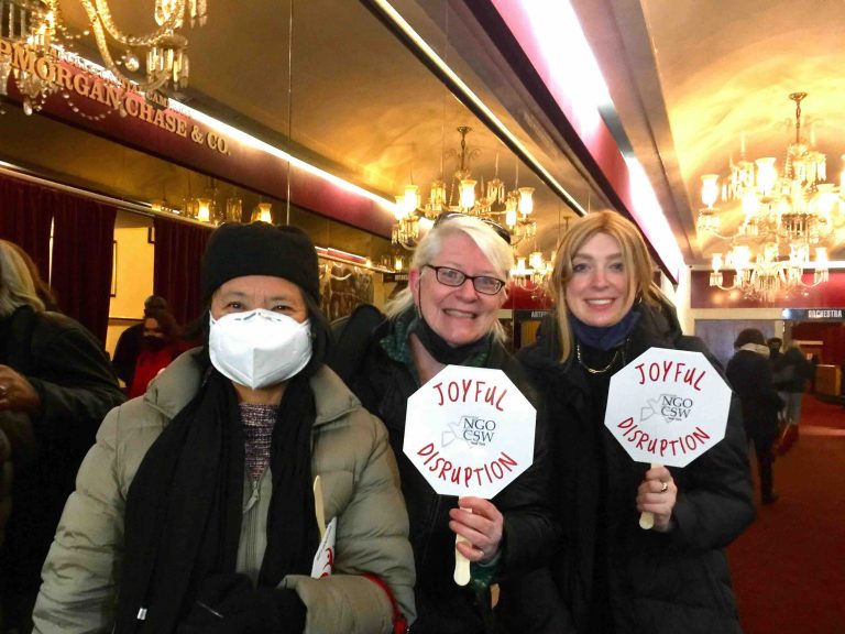 Three women, Janet Palifox, Beth Blissman, and Jax Viteznik, wearing thick winter jackets, and scarfs smiling together for a group picture indoors at a social event. One woman, Janet Palifox, is wearing a hat and a KN-95 mask, and the other two women, Beth Blissman and Jax Viteznik are holding small white octagon signs with a NGO CSW logo in the center and the large red words: Joyful Disruption, surrounding the logo.