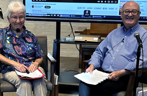 A man and woman sitting together with assorted mics and a display screen behind them. They are smiling for a picture with papers in their laps, likely preparing to present something with the display screen and microphones.