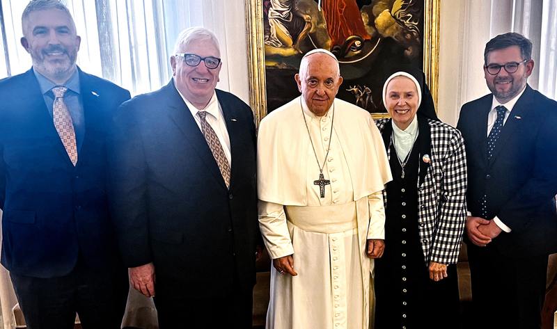 Three men wearing suits and ties, one nun, Jeannine Gramick, wearing a black and white plaid blazer, and a Pope, Pope Francis, in the center all smiling together for a group picture inside a church in front of a large detailed painting in a gold frame and large windows with white curtains on either side.