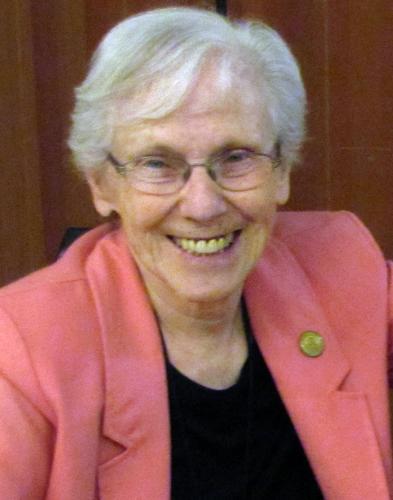 A woman with short white hair and wire glasses smiling brightly for a headshot picture wearing a black blouse and salmon pink blazer in front of a plain dark brown wall.