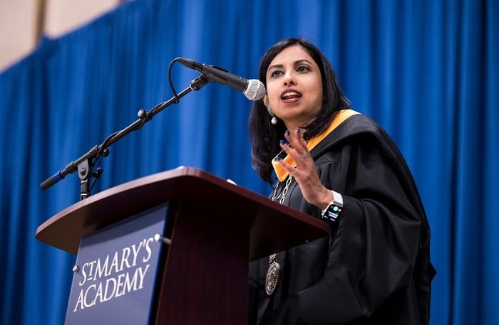 A woman, Iswari Nararajan, with straight brown hair, wearing a ceremonial black robe and yellow stole speaking passionately at a microphone on a wooden lectern with a dark blue banner with white text that says: St. Mary's Academy in front of large dark blue curtains.