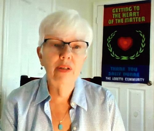 A woman with short white hair and glasses speaking to the camera while sitting in her office. Behind her is a strategically placed banner that is fully visible in the frame. Assumably relating to the online meeting she is attending when photographed.