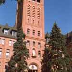 A red stone building featuring a tall tower above the arched entrance. The tower and entrance are framed by two large spruce trees that are only half as tall as the tower.