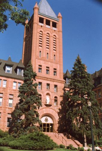 A red stone building featuring a tall tower above the arched entrance. The tower and entrance are framed by two large spruce trees that are only half as tall as the tower.