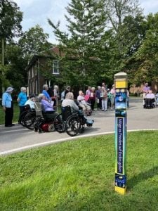 A colorful peace poll stands in the middle of a grassy area while people gather behind it on the pavement.