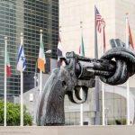 Large sculpture of a gun with the barrel tied in a knot, it is placed behind many flags of the world.