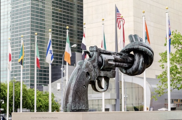 Large sculpture of a gun with the barrel tied in a knot, it is placed behind many flags of the world.