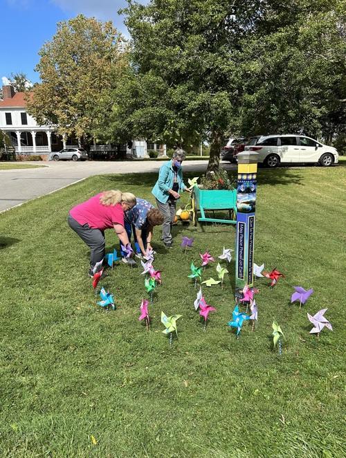 Three women work together to decorate a large patch of grass with handmade paper windmills. The field also has a bright teal wooden wheelbarrow with assorted flowers and a hand-painted pole surrounded by the paper windmills.