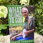 A book cover featuring a man with short grey hair and glasses playing a banjo and smiling with the title in bold white letters: What Belongs to God by David Edwards. The book is displayed standing on a rock in front of large white flowers and greenery.