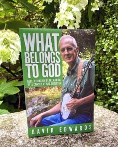 A book cover featuring a man with short grey hair and glasses playing a banjo and smiling with the title in bold white letters: What Belongs to God by David Edwards. The book is displayed standing on a rock in front of large white flowers and greenery.