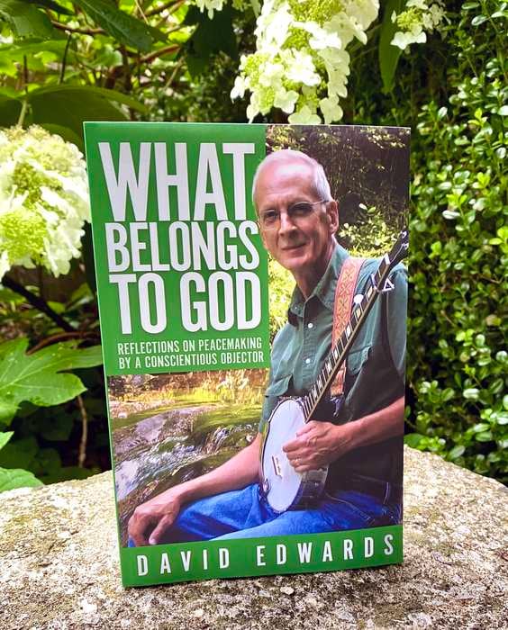 A book cover featuring a man with short grey hair and glasses playing a banjo and smiling with the title in bold white letters: What Belongs to God by David Edwards. The book is displayed standing on a rock in front of large white flowers and greenery.