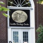 The front of a brick building with white french doors, an arched window, and the name plaque: Loretto Heritage Center featuring a vintage-inspired logo of a drawing of an old building. Stairs and a handrail are leading up to the doors and the photo features green leafy branches around the perimeter of the picture.