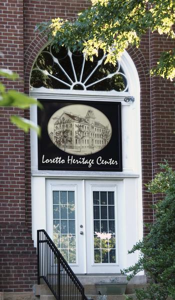 The front of a brick building with white french doors, an arched window, and the name plaque: Loretto Heritage Center featuring a vintage-inspired logo of a drawing of an old building. Stairs and a handrail are leading up to the doors and the photo features green leafy branches around the perimeter of the picture.