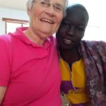 A woman, Mary Ann Gleason, with short grey hair, glasses, and a pink shirt smiling brightly while embracing her friend from Uganda indoors. The friend is a young female cancer patient with a shaved head wearing cultural yellow and purple clothing.