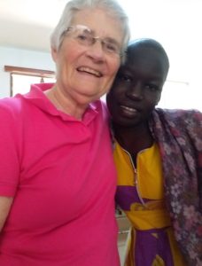 A woman, Mary Ann Gleason, with short grey hair, glasses, and a pink shirt smiling brightly while embracing her friend from Uganda indoors. The friend is a young female cancer patient with a shaved head wearing cultural yellow and purple clothing.
