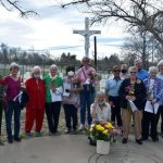 Fifteen people gathered at an outdoor cemetery with flowers smiling together for a picture on a sunny day in front of a large white crucifix statue.