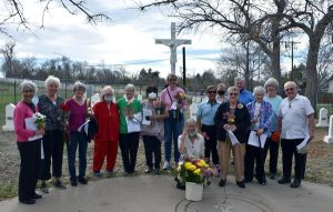 Fifteen people gathered at an outdoor cemetery with flowers smiling together for a picture on a sunny day in front of a large white crucifix statue.
