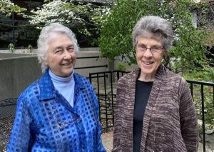 Two women, Jeanne Deuber and Marie Ego smiling together outdoors in front of a black metal fence with grass and trees past it. They both have short grey hair, Marie on the left is wearing a bright blue turtleneck and dark blue sweater and Jeanne on the right is wearing glasses, a black blouse, and a brown textured cardigan.