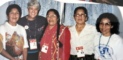 Five women smiling together for a group picture indoors, they are all wearing lanyard with large photo ID's.