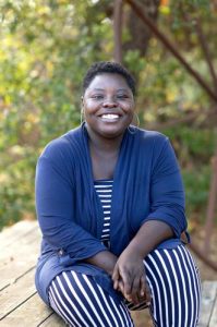 A woman, Tosha Phonix, with short brown hair smiling brightly for a portrait picture wearing big hoop earrings, a white and dark blue striped jumpsuit and a dark blue cardigan sitting outdoors on a wooden picnic table with trees and greenery in the background.