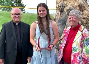A woman with long brown hair, wearing a blue dress with white detailing and assorted bracelets smiling with a man and woman in dress clothes while displaying a polished wood and glass trophy at an outdoor award ceremony on a sunny day.