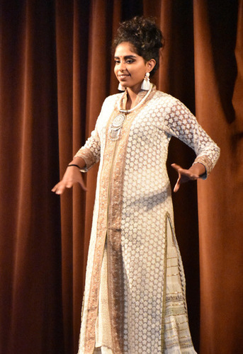 A woman wearing a white and gold cultural robe outfit from India. Her dark colored hair is pulled up in a bun, she has large earrings and a large necklace, and a bindi in between her eyebrows. She is raising her arms as if she is preforming while standing on stage in front of a brown velvet curtain.