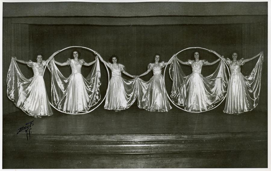 Teenage girls in shimmering dresses dance on stage with two large rings as props.