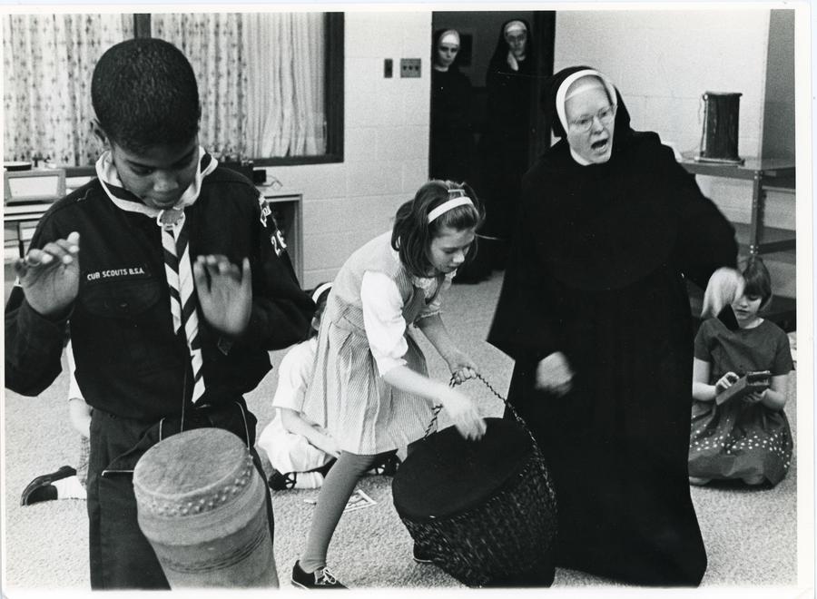 A nun in habit enthusiastically conducts a drumming class of older kids.
