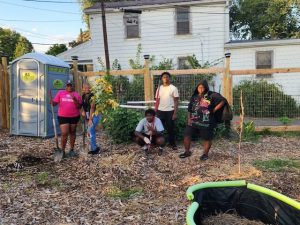 One woman in a pink shirt stands with four young people holding garden equipment standing next to a tree they planted in their garden.