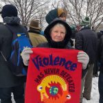 A woman in a heavy black jacket and wool gloves stands in a crowd with snow on the ground holding a red banner that says "violence has never defeated violence"